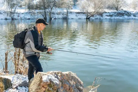 Mann angelt bei Minusgraden am Flussufer, das mit Schnee bedeckt ist. Er trägt eine Jacke und einen Rucksack.