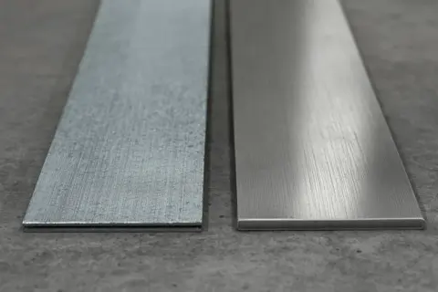 Dwa metalowe paski leżą obok siebie na betonowym podłożu. Jeden jest szorstki, drugi gładki, jakby przygotowany do montażu kotwy.
