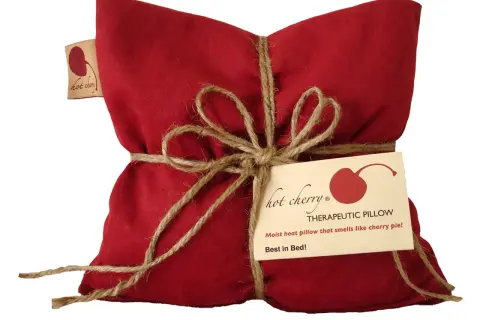 Cuscino rosso con noccioli di ciliegia, legato con spago. Un'etichetta recita "Hot Cherry Therapeutic Pillow". Benefici rilassanti.