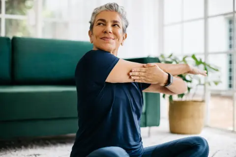 Une femme souriante s'étire, prouvant qu'il est possible de redresser son dos à 60 ans. Elle est assise sur un tapis, près d'un canapé vert.