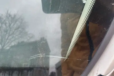 Głęboka rysa na szybie samochodowej, widoczna na tle rozmytego drzewa i budynku.