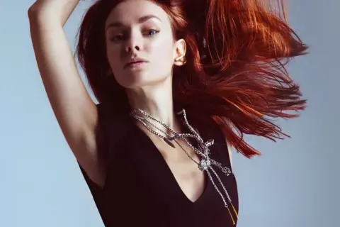 Mujer con cabello cobrizo vibrante, moviéndose con gracia. Luce un collar plateado llamativo y un pendiente dorado.