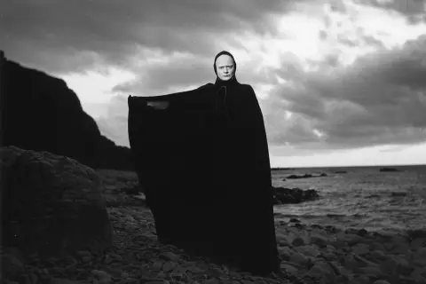 En skrämmande gestalt med vit ansiktsmålning och svart kåpa står på en stenig strand. En ikonisk scen från filmer med Ingmar Bergman.