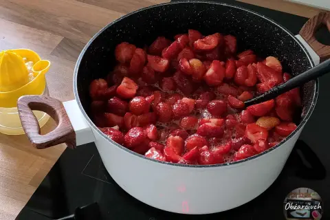 Duży garnek z gotującymi się truskawkami na kuchence indukcyjnej. Obok żółta wyciskarka do cytrusów.