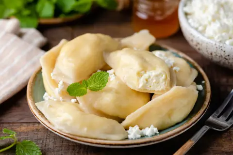 Pyszne pierogi z serem, udekorowane miętą i posypane serem. Obok słoiczek miodu i miseczka z serem.