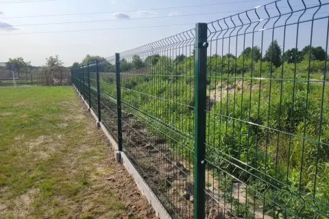 Nowe, zielone ogrodzenie działki rekreacyjnej z betonowym podmurowaniem. W tle trawa i zarośla.