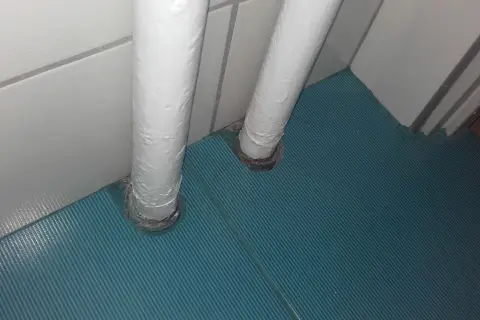 Dwa białe pionowe rury wychodzące z podłogi, przykryte zieloną wykładziną. Pomysł, jak zamaskować rury co przy podłodze.