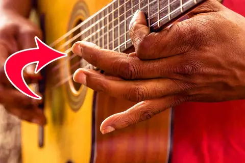 Nahaufnahme der Hände eines Fingerstyle-Gitarristen, der die Saiten einer Akustikgitarre spielt. Ein roter Pfeil zeigt auf die Finger.