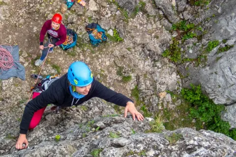 Wspinaczka podczas scala kursu. Kobieta w niebieskim kasku wspina się po skale, asekuruje ją osoba w czerwonym kasku.