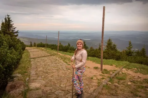 Uśmiechnięta turystka z kijkami na kamienistej ścieżce. Widok na góry i doliny. Czas wejścia na Śnieżkę zależy od kondycji i wybranej trasy.