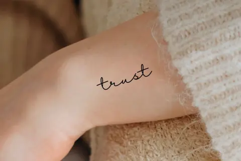 Ein zartes Tattoo mit dem Wort "trust" (Vertrauen) auf einem Arm, umgeben von weichem Strick.