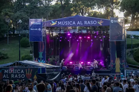 Escenario iluminado con luces púrpuras para un concierto de **música al raso**. Una multitud disfruta del espectáculo al aire libre.