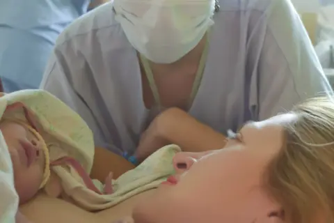 Noworodek przytulony do mamy, otoczony pieluchą. W tle widać personel medyczny w maseczkach, wspierający poród lotosowy.