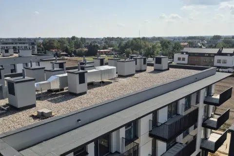 Widok na płaski dach z systemem wentylacyjnym i żwirem. Widoczne są warstwy stropodachu, elementy wentylacyjne i fragmenty budynków mieszkalnych.