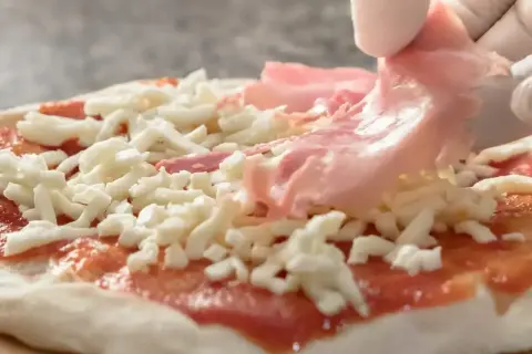 Rękawiczka w białej rękawiczce dodaje cienkie plastry różowej szynki na pizzę, która jest już pokryta sosem pomidorowym i startym serem.