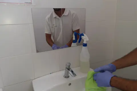 Man con guantes limpia lavabo con paño verde. Spray de limpieza cerca. Aprende como limpiar azulejos baño.