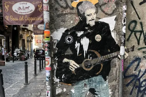 Graffiti di Pino Daniele con la chitarra, un omaggio a Napoli. I testi "Resta... resta cu'mmè" risuonano tra i vicoli.