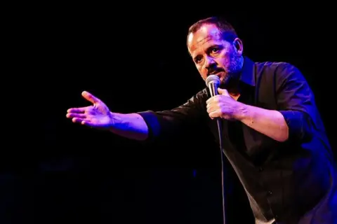Ángel Martín Zaragoza, comediante, habla al micrófono con gesto expresivo y mano extendida, iluminado por luces azules y púrpuras.