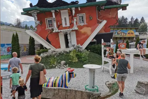 Ein rotes Haus steht auf dem Kopf, die Treppe führt ins Nichts. Eine Familie bestaunt das Spektakel, bunte Skulpturen und ein Dino-Land im Hintergrund.