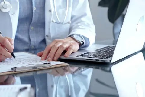 Lekarz rodzinny wypisuje receptę na depresję. Na biurku leży laptop i dokumenty.