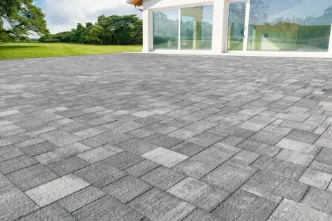 Un ampio patio in gres carrabile grigio, con pavimentazione a motivi geometrici, si estende davanti a una moderna casa con vetrate.