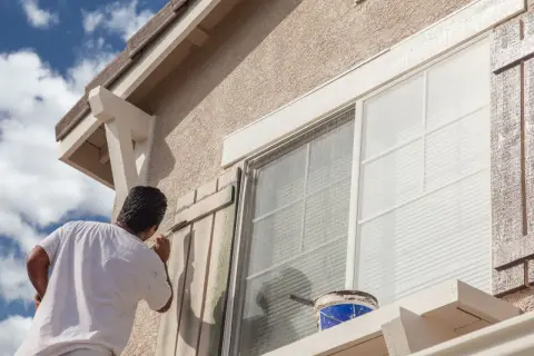 Hombre pintando persianas de madera para el acabado fachada de una casa. Cubo de pintura azul y blanca a la vista.