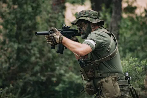 Jugador de deporte airsoft con camuflaje facial y equipo táctico, apuntando con su réplica de rifle en un bosque.