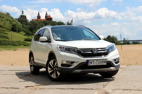 Biała Honda CR-V 2.0 benzyna na tle piaszczystego brzegu i zielonego wzgórza z budynkami.