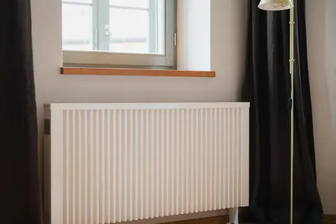 Biały ekran zagrzejnikowy stoi przy oknie, obok ciemnej zasłony i stojącej lampy. Idealny do wnętrz, gdzie liczy się estetyka i funkcjonalność.