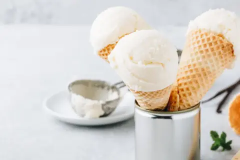 Cremiges griechischer joghurt eis ohne eismaschine in Waffelhörnchen. Ein Löffel und Vanilleschoten liegen daneben.