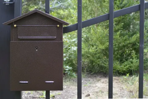 Brązowa skrzynka na listy w kształcie domku, zamontowana na czarnym słupku ogrodzenia. Zamykana na kluczyk, z miejscem na nazwisko.