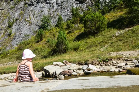 Dziecko w kapelusiku siedzi nad strumieniem w Małych Pieninach, otoczone zielenią i skalistym zboczem.