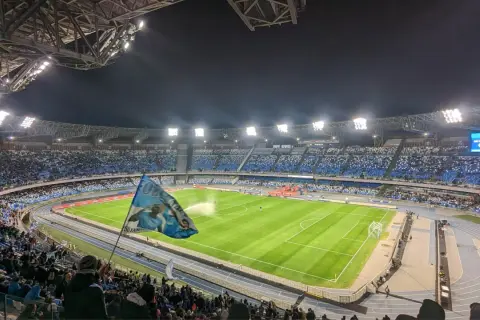 Widok na pełen kibiców stadion w Neapolu nocą. Trybuny wypełnione niebieskimi barwami, na murawie zielona trawa.