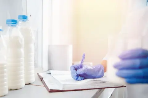 Controllo qualità del latte: un tecnico con guanti blu scrive appunti su un quaderno, accanto a bottiglie di latte.
