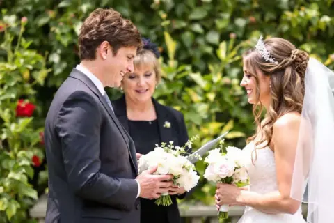 Cérémonie de mariage : une marraine émue lit une citation pour sa filleule rayonnante, entourées de fleurs.