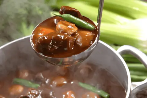 A ladle scoops tender, slow-cooked meat in rich gravy from a steaming pot. Perfect for learning jak dusić mięso.