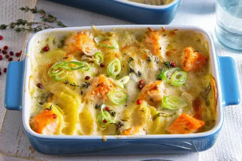 Herzhaft überbackener Lachs mit Kartoffeln in einer blauen Auflaufform, garniert mit Frühlingszwiebeln und rosa Pfefferbeeren.