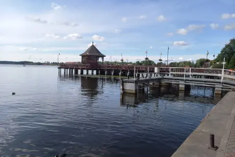 Drewniana altana na molo w Ostródzie, otoczona wodą i zielenią. Kaczki pływają spokojnie.