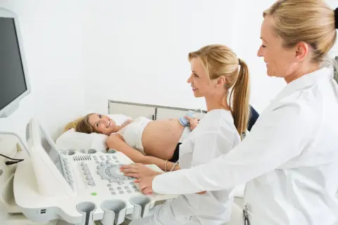 Lekarz ginekolog na NFZ wykonuje badanie USG ciężarnej pacjentki. Uśmiechnięta kobieta leży na kozetce, a lekarz obsługuje aparat.