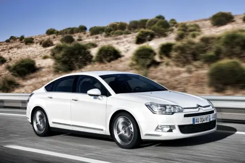 Biały sedan Citroën C5 w ruchu na drodze z pagórkowatym, zadrzewionym krajobrazem w tle.