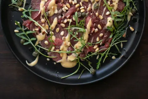 Rinder Carpaccio mit Pinienkernen, Kapern und Rucola auf schwarzer Platte, beträufelt mit einer cremigen Sauce.