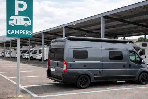 Estacionamiento para campers bajo techo. Un letrero verde indica "CAMPERS". Varios vehículos recreativos, incluyendo una furgoneta gris en primer plano, están aparcados.