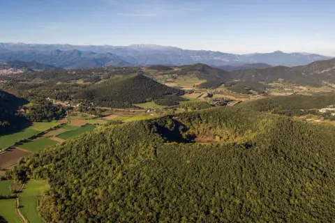 Paisaje de la ruta volcanes Garrotxa, con conos volcánicos cubiertos de bosque y campos verdes, bajo un cielo azul.
