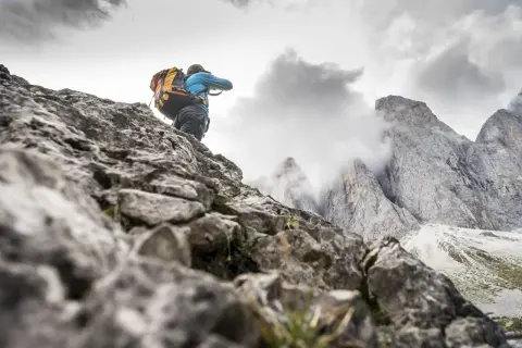 Un randonneur avec un grand sac à dos escalade un sentier rocheux, les pics des Dolomites se dressent dans le brouillard.