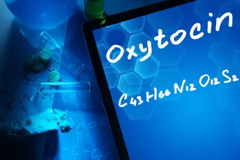 Na ekranie tabletu widnieje napis "Oksytocyna co to" i jej wzór chemiczny. W tle leżą tabletki, proszek i probówki.