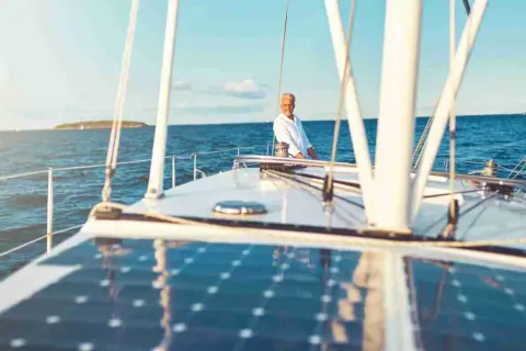 Un uomo sorride sul ponte di una barca a vela, con un impianto pannello solare che riflette il cielo azzurro.