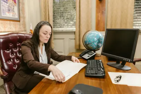 Kobieta w garniturze przegląda dokumenty, przygotowując się do wejścia w życie nowych przepisów. Na biurku globus, telefon i komputer.