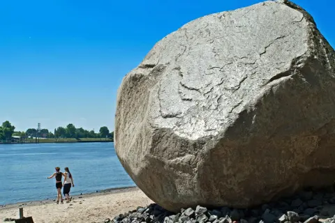 Der alte Schwede, ein riesiger Findling, thront am Ufer. Zwei Frauen spazieren am Wasser entlang, im Hintergrund Bäume und ein blauer Himmel.