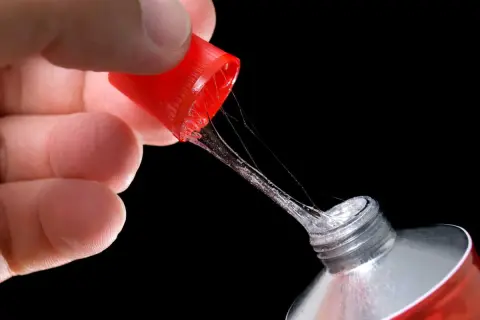 Main d'une personne tenant le bouchon rouge d'un tube de colle forte, avec des fils de colle s'étirant. Astuce pour comment enlever de la colle forte sur du métal.