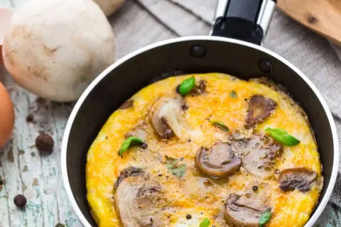 Pyszny omlet z pieczarkami, świeżo usmażony na patelni, z listkami bazylii i jajkami w tle.
